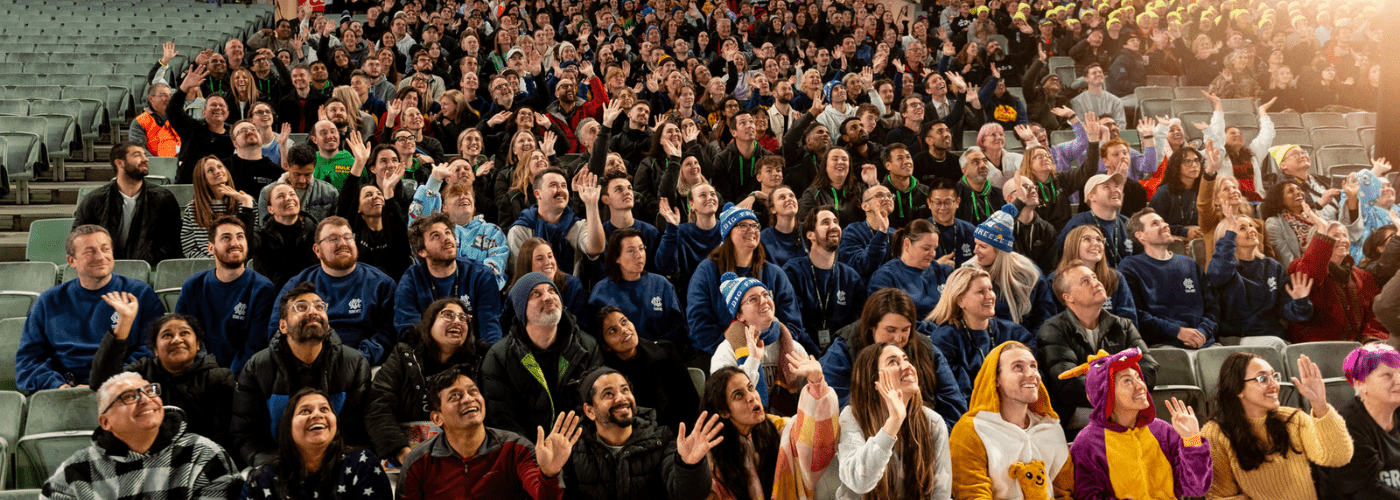 photo of a crowd of people in the stands of the MCG waving up at a drone camera